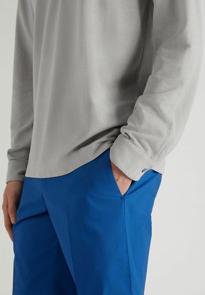 Grijze langwerpige shirt met een ronde zoom, gecombineerd met blauwe broek met een zijzak. Gladde stoftextuur, minimale detailing.