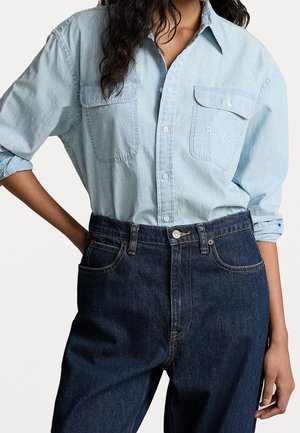 Personne portant une chemise en jean bleu clair à boutons avec les manches retroussées et un jean taille haute bleu foncé, debout avec une main sur la hanche.