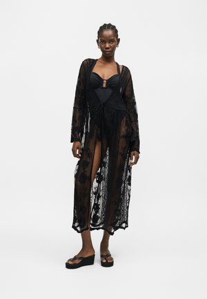 Žena v čiernych jednodielnych plavkách pod priehľadným dlhým čiernym čipkovým prekrytím, stojaca v čiernych klinových sandáloch na jednoduchom pozadí.