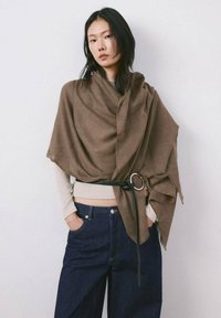 Mujer con cabello largo y oscuro que lleva un chal marrón asegurado con un cinturón negro sobre una blusa beige y jeans azul oscuro, de pie frente a una pared blanca.