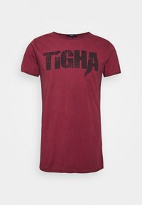Camiseta de manga corta burdeos con "TIGHA" en grandes letras negras desgastadas en el pecho, cuello redondo y ajuste casual.