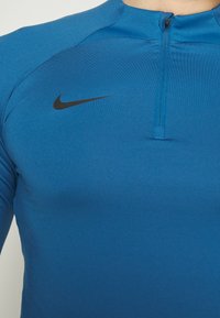 Maglietta sportiva a maniche lunghe blu con zip a un quarto. Presenta un logo Nike nero sul lato sinistro del petto. Tessuto liscio e elastico.