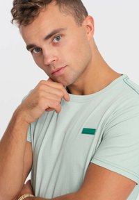 Světle zelené tričko s krátkými rukávy, kulatým výstřihem, kontrastním zeleným prošíváním a malou zelenou nášivkou na hrudi.