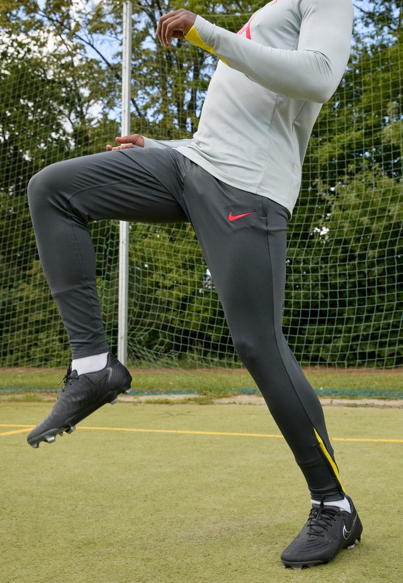 Pantalones deportivos grises con un acento amarillo, combinados con botas de fútbol negras. Material suave, diseño ajustado. Logo de Nike mostrado de manera destacada.