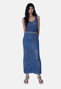 Robe deux pièces tricotée bleue avec un débardeur et une longue jupe, présentant un design en open weave, accentuée par une ceinture marron et des sandales blanches.