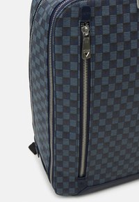Mochila azul-marinho com um padrão xadrez, tecido texturizado, dois fechos metálicos prateados e uma marcação discreta na lateral.
