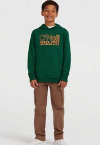 Felpa verde con testo arancione "O'Neill Santa Cruz California U.S.A.", tasca a marsupio, indossata con pantaloni cargo marroni e sneakers bianche.