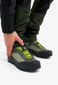 Person adjusting cuff of green and black hiking pants over green and black hiking shoes with yellow laces on white background.