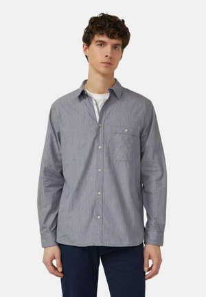 Junger Mann, der ein graues gemustertes Hemd über einem weißen T-Shirt und dunklen Hosen trägt, vor einem einfarbigen hellen Hintergrund stehend.