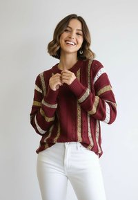 Bordowy sweter z dzianiny z pionowymi paskami w kolorze złotym i białym. Posiada ściągacze na mankietach i okrągły dekolt. Noszony z białymi spodniami.