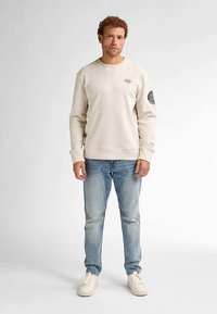 Lysebeige sweatshirt med rund halsudskæring og ribbede manchetter, kombineret med lyseblå jeans og offwhite sneakers. Har et broderet mærke.