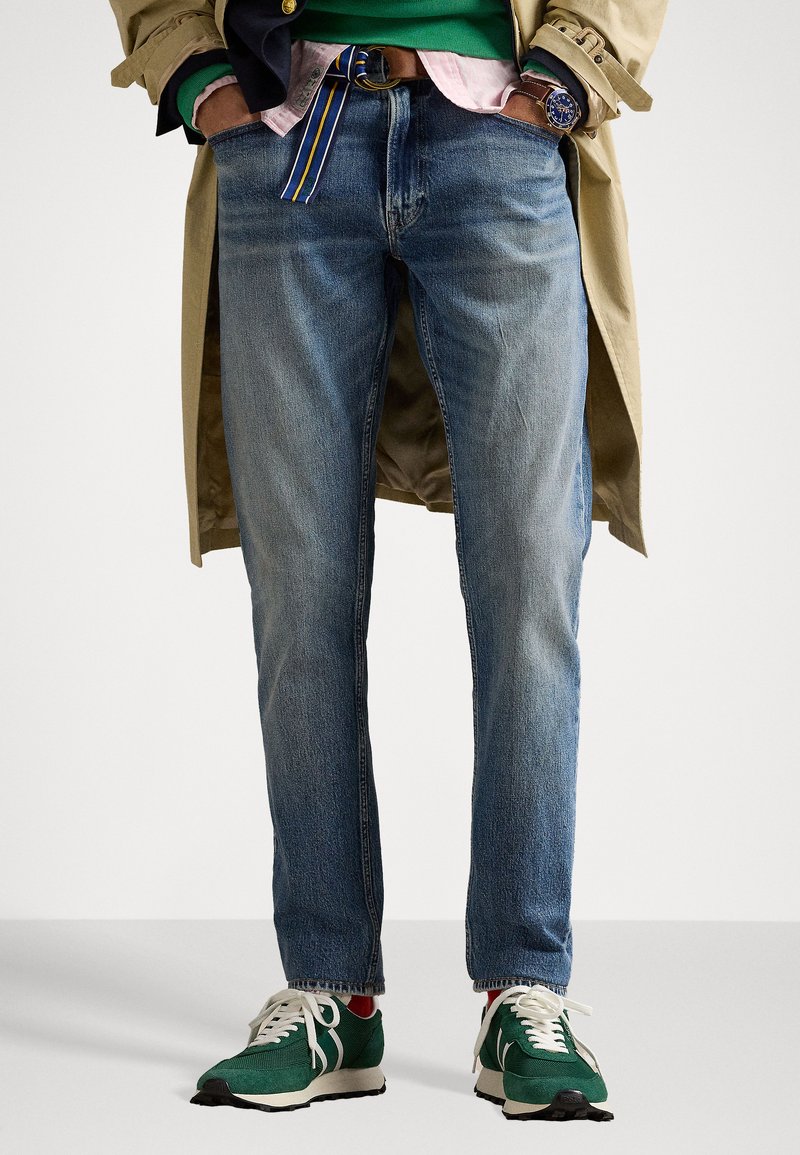 Jeans in denim blu con taglio dritto, abbinati a sneakers verdi. Presentano una cintura a strisce multicolori e dettagli di cucitura visibili.