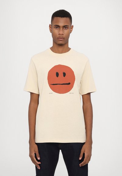 Beige Baumwoll-T-Shirt mit einem großen roten Smiley-Gesicht-Graphic, schwarzen Augen und Mund, runder Ausschnitt und kurzen Ärmeln.