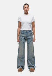 Jeans larghi blu chiaro con dettagli strappati, vita alta e tasche laterali, abbinati a una maglietta bianca aderente e calzature marroni.