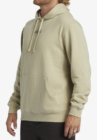 Sudadera beige con un bolsillo frontal, capucha con cordón, puños acanalados y un pequeño logo en el pecho. Material suave con una textura lisa.