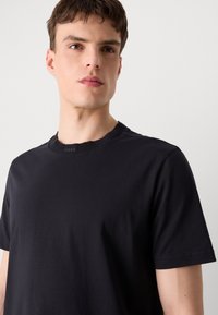 Μαύρο T-shirt από μαλακό βαμβάκι, με ραμμένο στρογγυλό λαιμό και κοντά μανίκια. Το λογότυπο της μάρκας είναι διακριτικά τοποθετημένο στον λαιμό.