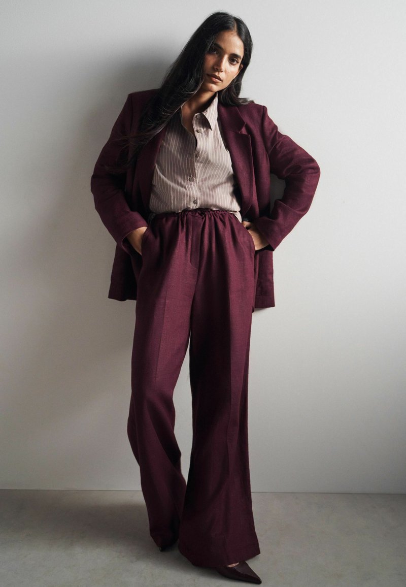 Naine seisab seina vastas, seljas burgundia värvi ülikond laia säärega pükstega, sobiva bleiseri, triibulise beežika särgi ja teravate ninaosadega kingadega.