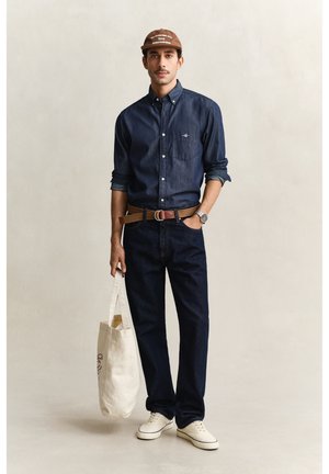 Mann trägt dunkles Jeanshemd und Jeans, braune Kappe, hellbrauner Gürtel, weiße Sneakers, hält eine weiße Tragetasche vor neutralem Hintergrund.