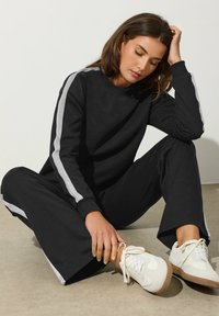 Donna seduta per terra, che indossa un completo sportivo nero abbinato con strisce grigie e scarpe da ginnastica bianche, mentre tiene una mano sulla testa.