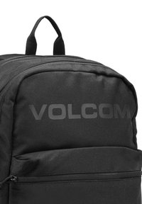 Black fabric backpack features a front zipper pocket, top carry handle, and the logo "VOLCOM" printed prominently on the front panel.