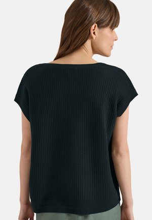 Femme aux cheveux bruns et raides portant un haut en maille texturée noire à manches courtes, vue de dos sur un fond uni.