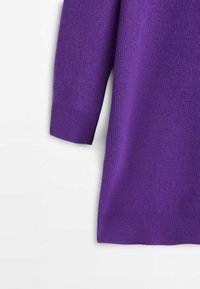 Détail de la manche longue et de la couture latérale d'un pull violet en tricot sur fond blanc.