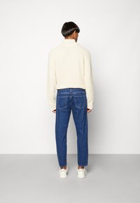 Bílý svetr s texturou a vysokým límcem, kombinovaný s modrými volně střiženými džíny a bílými teniskami, pohled zezadu.