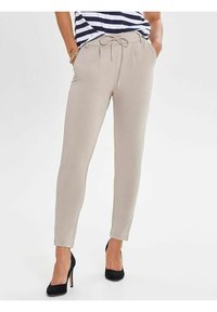 Pantalons beiges fuselés avec taille à cordon, portés avec des talons hauts noirs et un haut rayé, les mains dans les poches, pose debout.