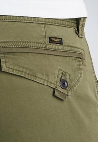 Olivgrüne Hosen aus strukturiertem Stoff, mit einer Klappentasche, die mit einem schwarzen Knopf gesichert ist, und einem schwarzen Etikett mit goldenen Flügel-Logo.