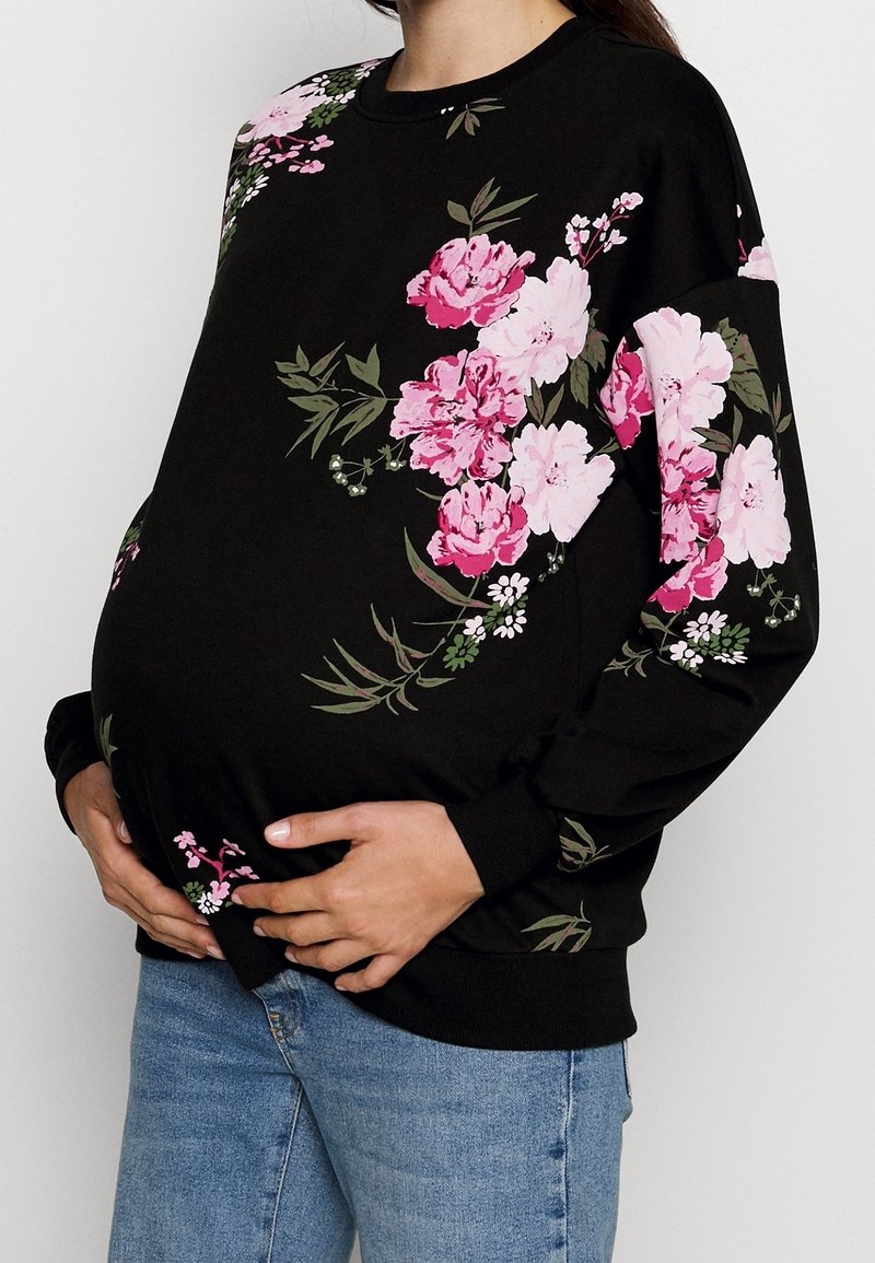 Sweat-shirt noir à motif floral rose, manches longues, col rond et poignets côtelés. Jean en denim clair porté. Mains posées sur le ventre.