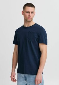 Tamsiai mėlynas medvilninis marškinėlis trumpomis rankovėmis, suapvalintu kaklu ir priekinėmis kišenėmis. Lygi tekstūra, laisvas siluetas, derinamas su šviesiai mėlynomis džinsinėmis kelnėmis.