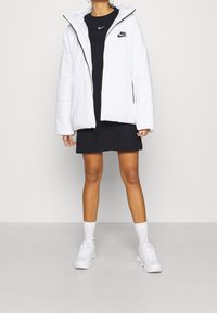 Vit puffjacka med huva, prydd med en svart Nike-logotyp. Bärs över en mörk klänning, matchad med vita sneakers och strumpor.