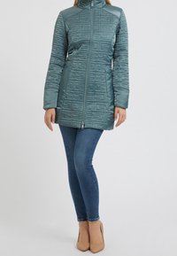 Tyrkysový péřový kabát s texturovaným geometrickým vzorem, vysokým límcem, plným zipem a dlouhými rukávy, kombinovaný s modrými džíny a světle béžovými podpatky.