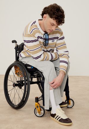 Νέος άνδρας σε αναπηρικό καροτσάκι προσαρμόζει γκρι φόρμα πάνω από λευκές κάλτσες και καφέ αθλητικά παπούτσια, φορώντας ριγωτό μακρυμάνικο πουκάμισο και γυαλιά ηλίου στο γιακά.