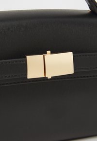 Sort håndtaske med en tekstureret overflade, der har en guldmetal-lås og et slankt design, kompletteret af en subtil stikning langs kanterne.
