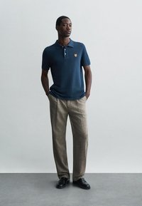 Vyras stovi su rankomis kišenėse, vilkintis tamsiai mėlynos spalvos polo marškiniais su mažu tigro logotipu ir smėlio spalvos plačiomis kelnėmis.