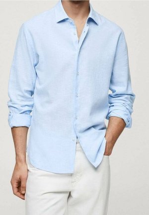 Mann trägt ein hellblaues, langärmliges Hemd mit aufgekrempelten Ärmeln, in weiße Hose gesteckt, eine Hand in der Tasche.