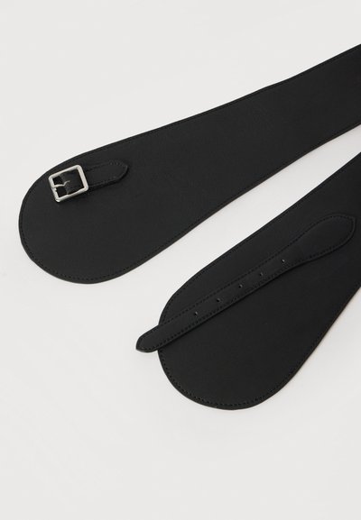 Μαύρες δερμάτινες λουρίδες με λείο υφή· ένα λουρί διαθέτει ασημένια μεταλλική αγκράφα και ρυθμιζόμενες τρύπες. Και τα δύο έχουν στρογγυλεμένα άκρα.