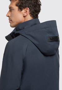 Chaqueta de marina con un alto cuello y capucha ajustable. Presenta un material texturizado y un parche con el logo en la parte posterior. Cierre de cremallera.
