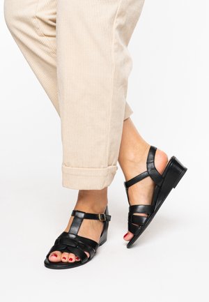 Schwarze Ledersandalen mit offenem Zehendesign, überkreuzten Riemen und niedrigem Absatz. Der Fuß trägt roten Nagellack.