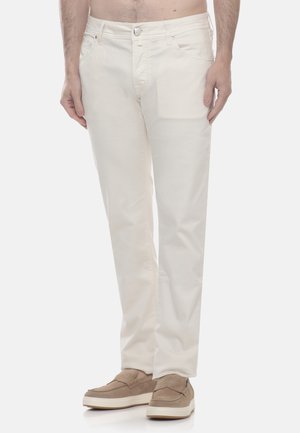 Modello maschile con pantaloni dritti color avorio e scarpe beige senza lacci, in piedi davanti a uno sfondo chiaro e uniforme.