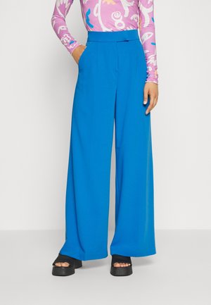 Osoba nosząca jasnoniebieskie szerokie spodnie z rękami w kieszeniach, w połączeniu z czarnymi sandałami na platformie i kolorową bluzką z długim rękawem.