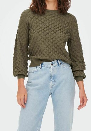 Pull olive vert avec un motif texturé en forme d'écailles, col rond et poignets côtelés ; associé à un jean taille haute bleu clair.