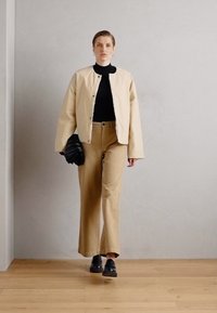 Beige bomberjakke med en svart genser med høy hals, brune vide bukser og svarte loafers. Holder en teksturert svart clutch. Jevn bakgrunn.