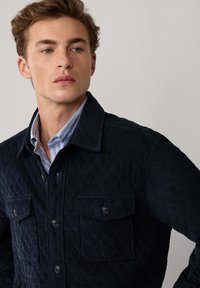 Granatowa pikowana kurtka z dwoma przednimi kieszeniami, zapięciem na guziki i kołnierzykiem. Teksturowana tkanina w diamentowy wzór. Jasnoniebieska koszula pod spodem.