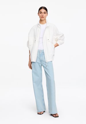 Femme portant une veste blanche zippée oversize, un haut blanc, un jean bleu clair à jambes larges et des talons ouverts noirs, debout devant un fond blanc uni.