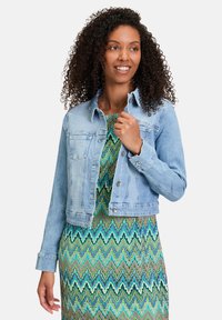 Veste en denim bleu clair avec fermeture à boutons, poches poitrine et manches longues. Portée au-dessus d'une robe verte à motif avec un design en zigzag.