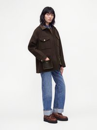 Жена, носеща тъмнокафяво oversized яке с големи джобове, къси сини дънки и кафяви кожени обувки, стояща пред бял фон.