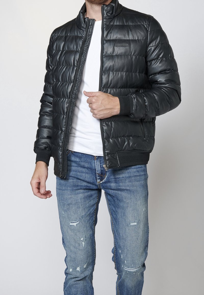 Homme portant une doudoune noire par-dessus un t-shirt blanc et un jean bleu usé, ajustant la fermeture éclair de sa veste avec la main droite.