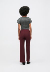 Mulher com cabelo afro a usar uma blusa metálica cinza de mangas curtas e calças largas em bordô, de pé, voltada de costas.